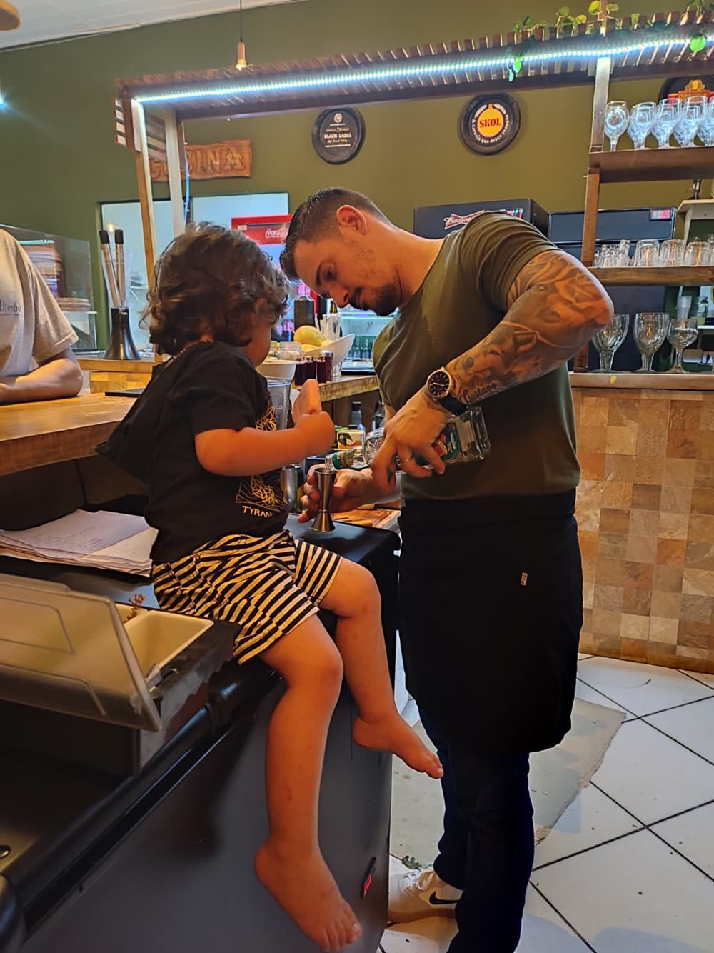 Renato no balcão da pizzaria com uma criança sentada ao lado