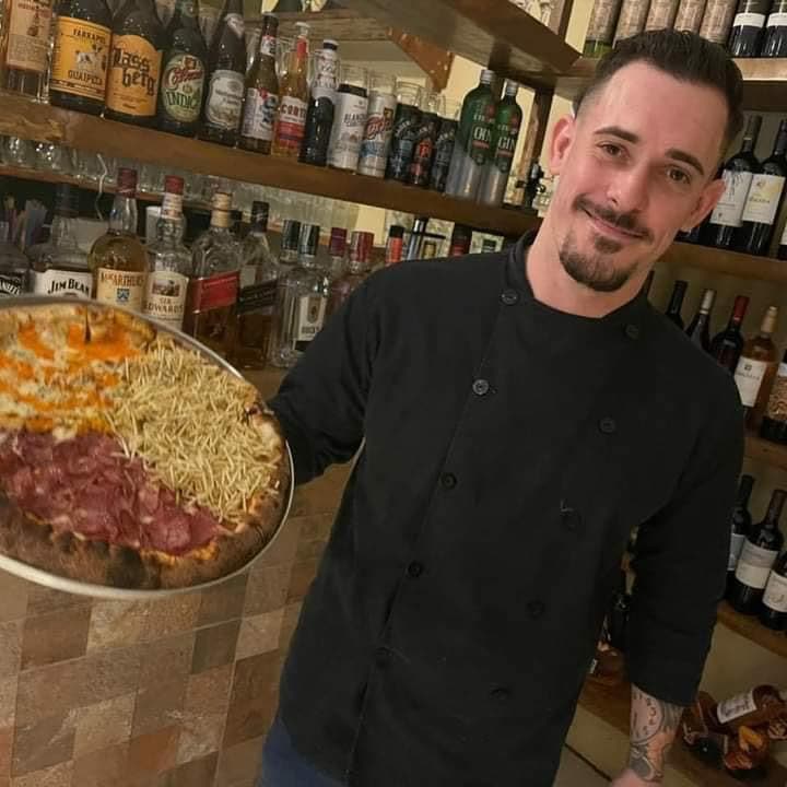 Renato na pizzaria, de dólmã preta, segurando uma pizza