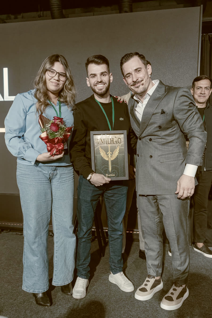 Aluna e aluno com a placa, ao lado do Renato no palco